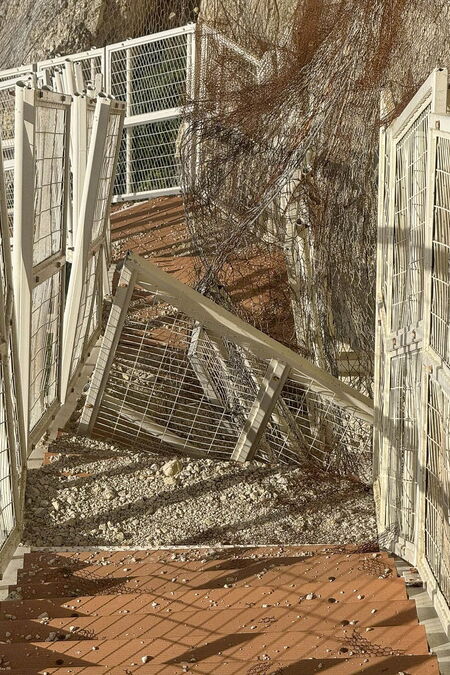 Eine in eine Klippe eingebaute Metalltreppe ist schwer beschädigt. Der weiße Metallzaun ist verbogen und über den orangefarbenen Stufen zusammengebrochen, die mit kleinen Steinen und Sand bedeckt sind.